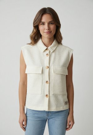 Femme portant un gilet sans manches blanc cassé avec deux grandes poches avant et un jean bleu clair, sur un fond uni.