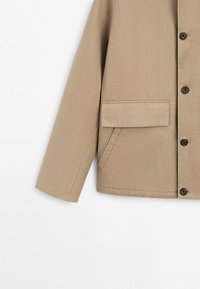 Beige langærmet jakke med knaplukning foran og klaplomme over den skrå sidelomme på den nederste højre forside.