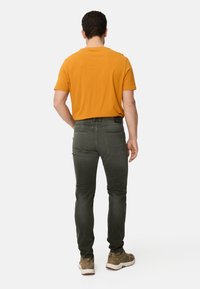 Orange kortærmet skjorte, mørkegrønne slim-fit jeans og brune sneakers med hvide såler. Bemærkelsesværdige syninger og lommedetaljer er synlige.