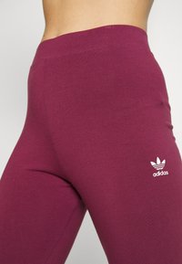 Bordó legíny vyrobené z měkkého a elastického materiálu. Má široký pas a bílé logo Adidas blízko dolní části nohavice.