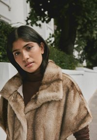 Faux fur outerwear in warm brown tones, featuring a wide collar and soft texture, layered over a fitted dark brown turtleneck.