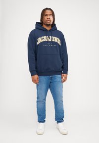 Mørkeblå hættetrøje med "JACK & JONES" logo i gul, med en frontlomme og ribbede manchetter. Båret med lyseblå jeans og hvide sko.
