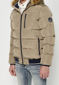 Homme portant une veste beige matelassée avec capuche bordée de fourrure, poches zippées, poignets côtelés bleu marine et jean bleu délavé sur un fond uni.
