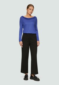 Langarmige blauwe top met gedrapeerde halslijn, gecombineerd met hoge, wijde zwarte broek en zwarte chunky loafers. Gladde, casual stof.