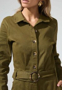 Robe boutonnée vert olive avec un col et une ceinture circulaire marron. Elle présente un tissu texturé et de grands boutons contrastants.