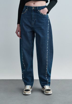 Sötétkék, bőszárú farmert viselő személy, ezüst szegecsekkel az oldalak mentén és a zsebeknél, bézs-fekete sportcipővel párosítva.