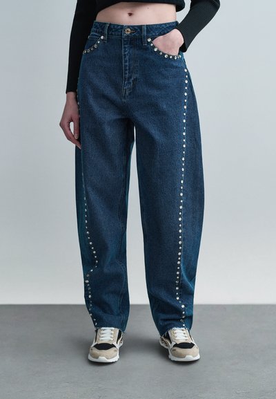 Personne portant un jean large bleu foncé avec des clous argentés le long des côtés et des poches, associée à des baskets beige et noires.