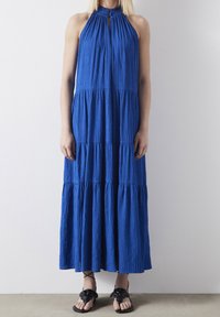 Robe maxi bleue sans manches en tissu texturé, avec un col froncé et un design à étages. Sandales noires avec des accents floraux.
