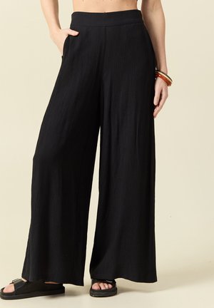 Persona che indossa pantaloni neri larghi e morbidi con tasche laterali, sandali neri con zeppa e braccialetti a un polso.