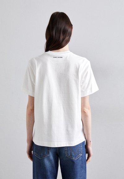 White cotton T-shirt with short sleeves, featuring a black "MARC JACOBS" logo on the upper back, paired with blue denim jeans.