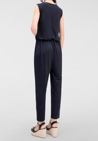 Marinblå jumpsuit med ärmlös design och elastiskt midjeband. Slät tyg med avslappnad passform, kombinerat med bruna kilklackar.