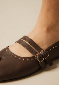 Chaussure Mary Jane en cuir marron avec deux brides à boucle et petits clous dorés sur le bord, portée sur un pied nu.