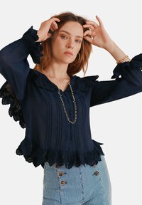 Navyblaue Bluse mit Spitzenakzenten, ausgestellten Ärmeln und strukturierten Falten. Besitzt einen Rüschensaum und einen knopfbaren Rock mit horizontalen Streifen.