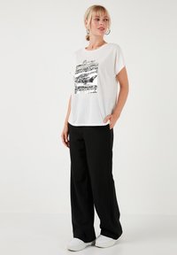 Hvid kortærmet t-shirt med et sort og sølv grafisk design, parret med sorte brede bukser og hvide sneakers.