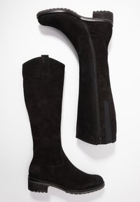 Botas altas de ante negro, con punta redonda, cremallera lateral y suela de goma texturizada. Cuentan con un tacón plano y una tirador decorativo.