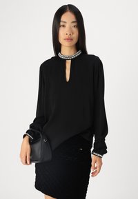 Blouse noire à manches longues avec un encolure en trou de serrure, ornée de poignets embellis. Portée avec une jupe noire texturée et un petit sac à main.