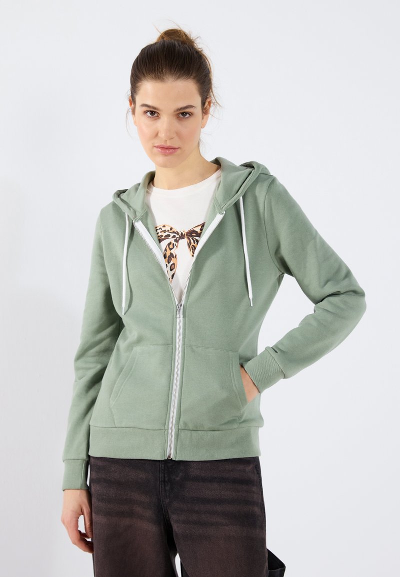 Menta zelena zip-up dukserica napravljena od mekanog materijala, sa prednjim zatvaračem, kapuljačom sa vezicama i prednjim džepovima. Nošena preko belog majice sa printom.