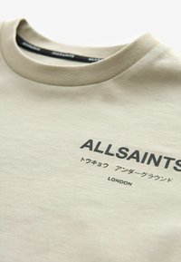 Beige puuvillane T-särk, millel on ümmargune kaelus, trükitud "ALLSAINTS" logo ja lisatekstid mitu keelt esiküljel.