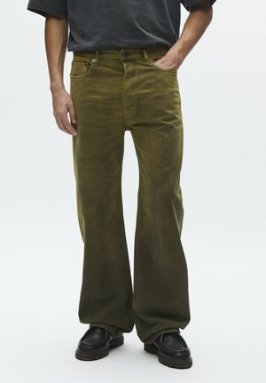 Homme portant un pantalon ample vert olive avec une chemise noire, les mains près des poches, et des mocassins noirs sur un fond blanc.