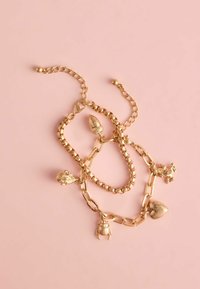 Guld charm-armbånd med teksturerede led, dekorative charms inklusive en akorn, hjerte, frø og bille, på en lyserød baggrund.