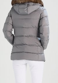 Giacca a vento grigia con trapuntatura orizzontale, cappuccio attaccato e un accento in pelliccia marrone rimovibile lungo il collo. Materiale liscio e lucido.