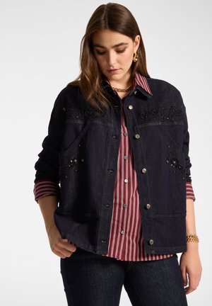 Jeune femme portant une veste en jean foncée avec une broderie noire sur une chemise à rayures rouges et blanches à boutons, sur fond uni.