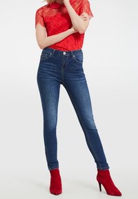 Top de encaje rojo, jeans azul de tiro alto con un ligero estiramiento, con diseño clásico de cinco bolsillos. Combinados con botas de tacón alto de ante rojo.