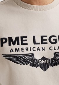 Svetle bež pulover s črnim vezenim napisom "PME LEGEND AMERICAN CLASS" in podrobno krilato emblemo spodaj. Rebrast ovratnik.