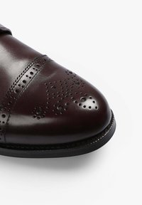 Zapato de cuero marrón con puntera redonda, perforaciones decorativas y una costura contrastante a lo largo de la parte superior. Suela de goma negra.