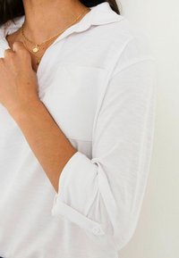 Weißes, langärmliges Hemd mit Kragen, ausgestattet mit einer Brusttasche. Weicher Stoff und hochgerollte Ärmel, die mit Knöpfen gesichert sind, sowie einem auffälligen Halsgeschmeide.