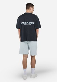 Čierne voľné tričko s logom "PEGADOR" v svetlom modrom odtieni na zadnej strane, doladené so svetlomodrými rebrovanými šortkami a bielymi teniskami.