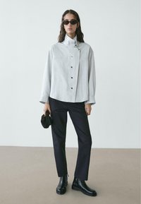 Camicia oversize grigio chiaro con collo alto, abbinata a pantaloni neri e stivaletti alla caviglia. La modella tiene una piccola borsa nera.