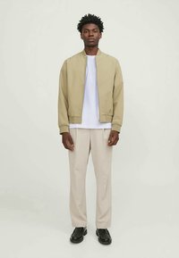 Young man standing wearing a beige bomber jacket, white t-shirt, light beige trousers, and black shoes against a plain light background.