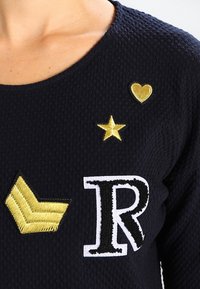 Tmavě modrý texturovaný vršek s designem ze zlatých vyšívaných akcentů: srdce, hvězda a písmeno "R", s detailním vzorem šipky na rukávu.