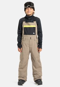 Salopettes de ski beiges avec un panneau en tissu noir sur la poitrine et une poche zippée jaune. Tissu texturé, bretelles ajustables et zips latéraux. Sous-vêtements de base noirs et lunettes de ski.
