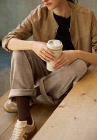 Hnědý bomber kabát, černý top a béžové kordové kalhoty. Drží texturovaný bílý hrníček na kávu. Hnědé ponožky a světlé tenisky s tmavými akcenty.