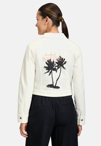 Veste en denim blanc avec un graphique de palmier noir et le texte "beauté de la nature" en accent métallique. Elle possède un col et des poignets boutonnés.