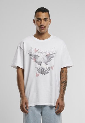Homme aux cheveux courts et moustachu porte un t-shirt blanc avec un grand dessin de colombe blanche et grise et des petits oiseaux roses, jean bleu clair.