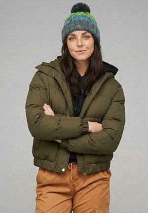 Woman with dark hair wearing green and blue knit hat, olive puffer jacket, black hoodie, and tan pants standing with arms crossed.