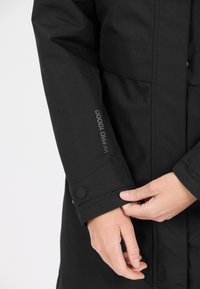 Svart vattentät jacka med texturerad tyg, justerbar mudd med tryckknapp och "W-PRO 10000"-logotyp på ärmen.