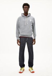 Junger Mann in grauem Kapuzenpullover, dunkler Hose und gelben Turnschuhen vor weißem Hintergrund.