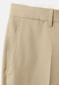 Beige bomuldsbukser tæt på, der viser linning, bæltestrop, sidelomme og detaljer ved forkanten.