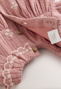 Vestito di cotone rosa con tessuto strutturato, ricamo floreale bianco, bordatura smerlata e bottoni di legno. L'etichetta recita "NEXT baby, made in India."