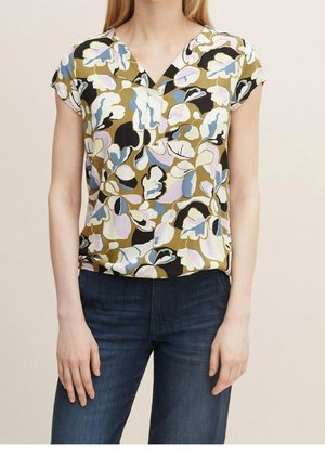 Kobieta ubrana w krótki rękaw bluzki w kwiatowy wzór w stonowanych zielonych, niebieskich, czarnych i białych kolorach, połączona z ciemnoniebieskimi dżinsami.