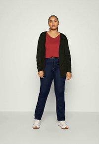 Svart cardigan, burgunder topp, mørkeblå jeans, hvite sneakers. Modellen står oppreist og viser frem et avslappet antrekk med strukturelt stoff.