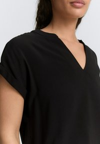 Schwarzes Kurzarmoberteil mit V-Ausschnitt, das aus einem eleganten Stoff mit glatter Textur gefertigt ist. Mit hochgerollten Ärmeln und einem lockeren Schnitt.