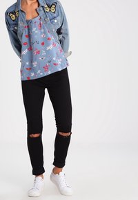 Blå blommig ärm-lös topp under en denimjacka med fjärilsapplikationer, ihop med svarta rivna jeans och vita sneakers.