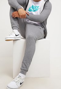 Grey cotton tracksuit with fitted joggers and a matching hoodie. Worn with a white t-shirt featuring a turquoise Nike logo and white sneakers.