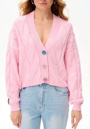 Kobieta ubrana w różowy sweter rozpinany z warkoczowym wzorem z trzema guzikami w różnych kolorach, na jasnoniebieskich dżinsach, na białym tle.