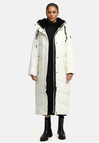 Long white puffer coat with black faux fur-lined hood, front zip, side pockets, and quilted design. Worn over a black ribbed dress and boots.